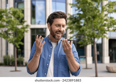 Close-up photo of a smiling young man standing on a city street talking on the phone through a loudspeaker, recording a voice message. - Powered by Shutterstock