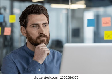 Close-up photo of a thoughtful and serious young man sitting in the office at the workplace in front of the laptop, thoughtfully looking ahead, holding his head with his hand. - Powered by Shutterstock