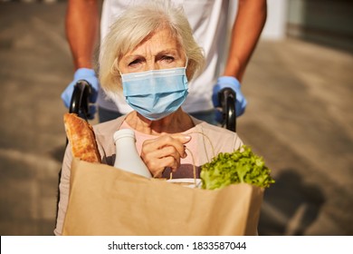 Close-up photo of a troubled aging person with a face mask keeping a brown bag with baguette, milk and lettuce - Shutterstock ID 1833587044
