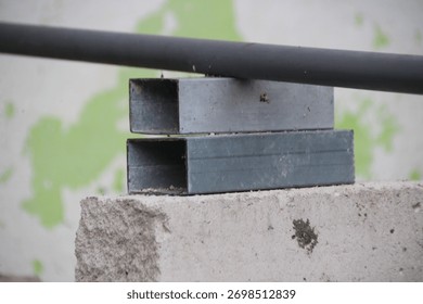 A close-up photo of two gray rectangular hollow metal bars (light steel) stacked on top of a rough concrete block. A dark round pipe rests on top of the stack of bars. The background is a blurry wall 