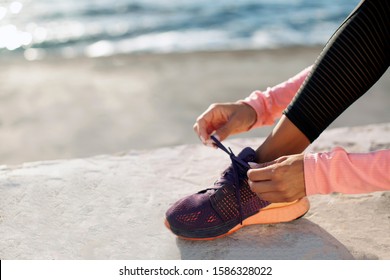 Closeup Photo Of Woman Lacing Her Shoes During Outdoor Workout By The Sea, Wearing Black Sport Pants And Pink Long Sleeve Shirt