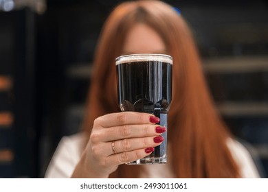 Close-up photo of a woman with red nail polish holding a glass of dark beer with a creamy head. The background is blurred, focusing on the beer and the woman's hand - Powered by Shutterstock