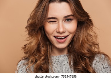 Close-up photo of young charming brunette woman blinks one eye and looking at camera, isolated over beige background