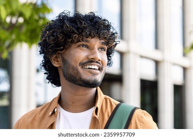 Close-up photo of a young Indian smiling man looking to the side with a confident look. Standing outside on the street with a backpack. - Powered by Shutterstock