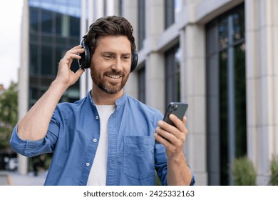 Close-up photo of a young man wearing headphones and using a mobile phone. Standing smiling in casual clothes outside on a city street. - Powered by Shutterstock