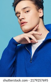Close-up photo. a young and slender man with thick black hair standing on a blue background in a blue zip-up sweater with a white T-shirt under it.