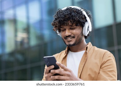 Close-up photo. Young smiling Indian male student standing outside wearing white headphones and holding phone. Calls, chats, listens to music.