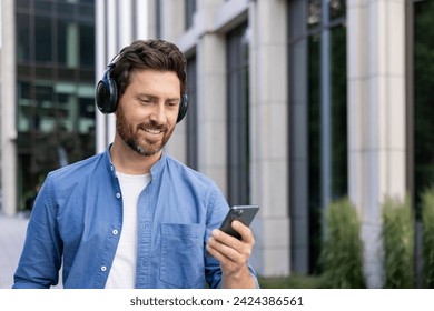 Close-up photo of a young smiling man standing on a city street wearing headphones and using a mobile phone. Listens to music, watches videos, talks on a video call. - Powered by Shutterstock