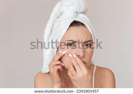 close-up photo of a young woman in white underwear with a white towel wrapped around her head, she looking at the camera, squeezing out a red pimple on her cheek with both hands and frowning. 