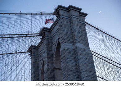 Close-up photograph of the Brooklyn Bridge tower, highlighting its stone architecture, suspension cables, and an American flag waving at the top.  - Powered by Shutterstock