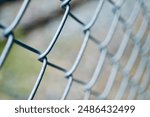 A close-up photograph of a chain-link fence. The image focuses on the intertwined metal wires, showcasing the texture and structure of the fence. The background is blurred, emphasizing the detail