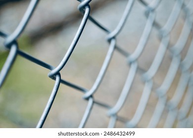 A close-up photograph of a chain-link fence. The image focuses on the intertwined metal wires, showcasing the texture and structure of the fence. The background is blurred, emphasizing the detail