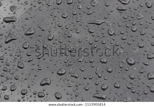 Closeup photograph of raindrops on\
the hood of a black car. Rain clouds are reflected in the\
hood.
