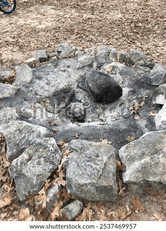 A close-up photograph of a rustic fire pit made of large stones, filled with ash and partially burned logs, surrounded by fallen autumn leaves. The fire has burned out, leaving behind remnants of a pa