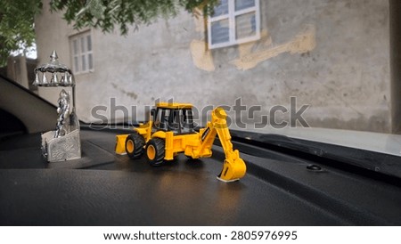 Close-up photograph of a silver Ramapir (Ramdev Pir) idol with a yellow toy backhoe loader placed on a car dashboard. Natural daylight with selective focus and shallow depth of field.