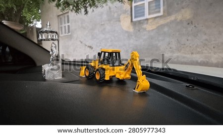 Close-up photograph of a silver Ramapir (Ramdev Pir) idol with a yellow toy backhoe loader placed on a car dashboard. Natural daylight with selective focus and shallow depth of field.