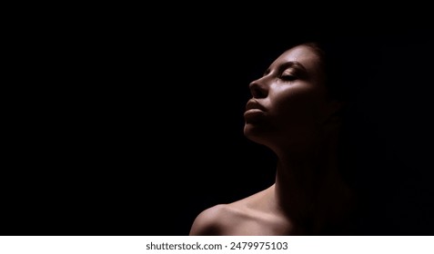 A close-up photograph of a womans face, captured in a dark environment. The woman is facing upwards, and her face is illuminated by a single light source, casting shadows across her features. - Powered by Shutterstock