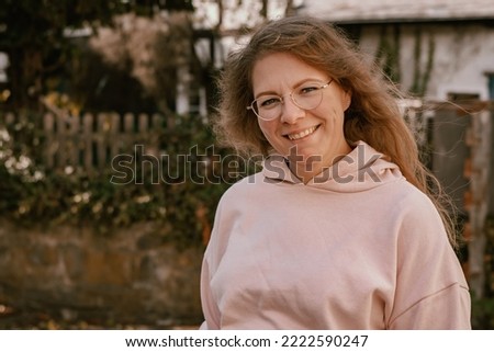 Close-up photography of a 30 years old woman showing a cute smile and looking straight into the camera; the woman is wearing a pink hoodie; the picture was taken outdoors in countryside