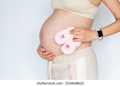 Closeup Photography Of Young Woman Holding Tiny Pink Shoes For Baby Daughter And Touching Hands Her Naked Big Belly Isolated On White Background With Copy Space. Skin Care With Moisturising Cream