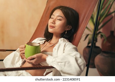Close-up picture of beautiful korean woman having rest after work enjoying her relaxing herbal tea and silence, sitting on lounger with closed eyes