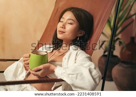 Close-up picture of beautiful korean woman having rest after work enjoying her relaxing herbal tea and silence, sitting on lounger with closed eyes