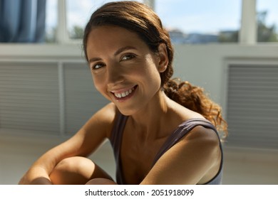 Close-up Picture Of Beautiful Woman Face With Mimic Wrinkles And Natural Nude Make-up, Posing Against Wide Window With Toothy Smile, Feeling Good Sitting On Floor Under Sun Rays In Sportswear