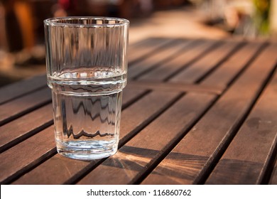 Close-up Picture Of A Glass Of Water Which Is Half-full Standing On A Brown Wooden Table And As The Sun Shines Through The Glass The Reflection Can Be Seen On The Table