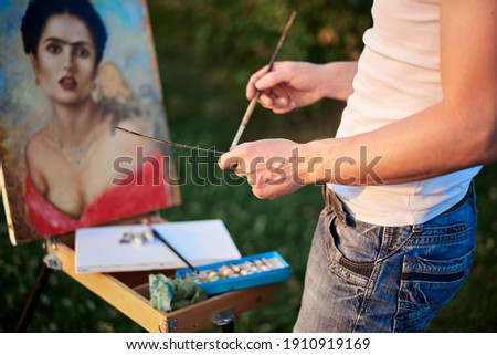 Close-up picture of hands of male artist, drawing female portrait on canvas on green field in summer. Painting workshop in rural countryside. Artistic education concept. Outdoors leisure activities.