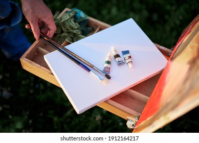 Close-up Picture Of Painting Equipment, Including Sketchbook Easel, Watercolor Tubes, Paint Brushes And Canvas. Arts Workshop Outside In Summer. Artistic Education Concept.