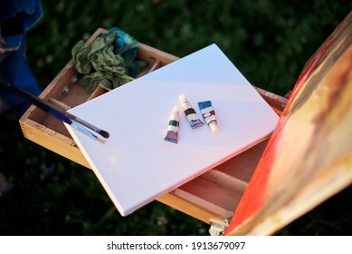 Close-up Picture Of Painting Equipment, Including Sketchbook Easel, Watercolor Tubes, Paint Brushes And Canvas. Arts Workshop Outside In Summer. Artistic Education Concept.