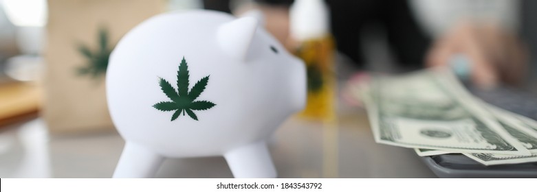 Close-up of piggy bank with green sign of cannabis. Paper package and bottle of cannabinoid oil with marijuana symbol on table. Cash on desk. Painkillers and medical marihuana concept
