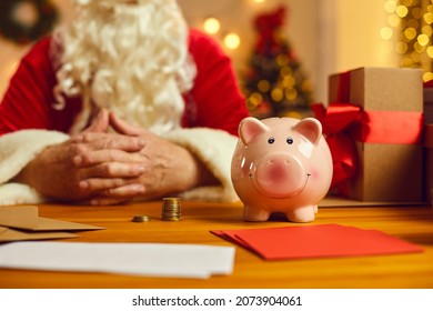 Close-up of piggy bank on desk with presents and Christmas charity event invitations with old Santa Claus in blurred background. Donation to orphan funds, saving up money, opening bank account concept