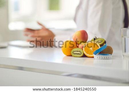 Close-up of pile of fresh apples, kiwis and oranges and measuring tape on office desk against blurred dietitian or nutritionist giving weight loss advice during consultation with patient
