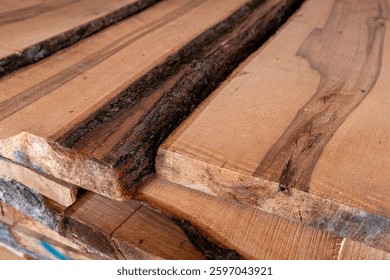 Close-up of piled stack of natural brown uneven rough wooden boards lit by bright sun. Sawmill. Wooden planks at a sawmill or in a carpentry workshop. Raw wood drying in the lumber warehouse