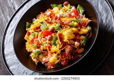 close-up of pinto beans bacon cornbread salad with tomatoes, peppers, corn and cheddar cheese in black bowl on dark wood table, horizontal view from above