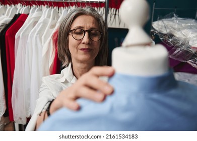 Close-up Pleasant Elderly White-haired European Female Tailor Working On New Clothes In Fashion Clothing Design Atelier, Putting A Shirt From Her Luxury New Collection On A Tailoring Mannequin
