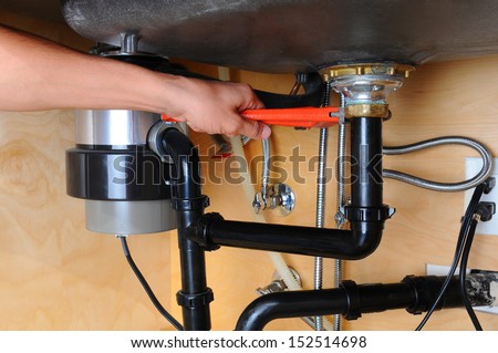 Closeup of a plumber using a wrench to tighten a fitting beneath a kitchen sink. Only the man's hand and arm are visible. Horizontal format.