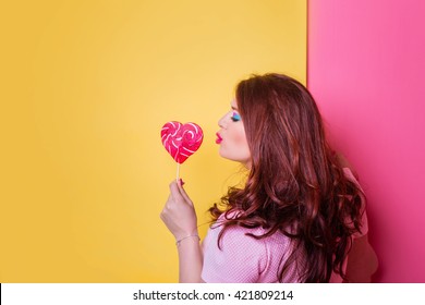 Close-up Plump Fat Plus Size Redhead Woman With Long Hair, Curly With Bright Make Up And Pink Lips With Sweet Heart Candy In Hand Handmade. Vector Yellow Pink Background. Unusual Emotion