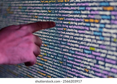 Close-up of a pointing hand over a large, glowing display screen filled with colorful code, possibly representing computer programming or data visualization