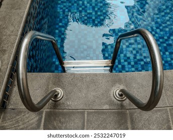 Close-up of pool stairs with metal handrails extending from the tiled pool deck into the water. Blue pool tiles. - Powered by Shutterstock