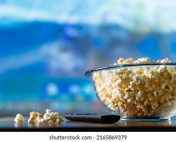 Close-up. Popcorn In A Bowl On A Blue Background And A TV Remote Control. There Are No People In The Photo. There Is Free Space To Insert. Home Comfort, Traditional Food For Watching Shows.