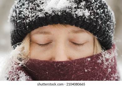 Close-up portait of a blonde woman in beanie hat and nose covered in scarf stands with eyes closed in winter snowfall. Nordic winter seasonal portrait, tranquil and calm outdoor scene