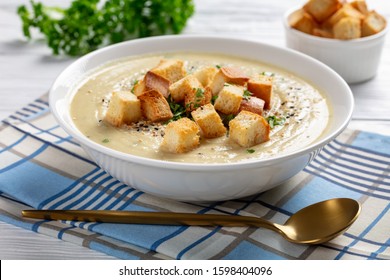 Close-up Of A Portion Of Asiago Roasted Garlic Cauliflower Soup Topped With Croutons In A White Bowl On A Wooden Table, View From Above,