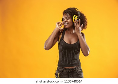 Close-up portrait of adorable curly girl happy smiling during photoshoot. Stunning african woman with light-brown skin relaxing in headphones and funny dancing on colorful yellow background at studio.