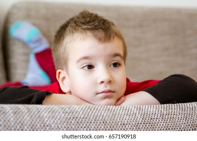 Close-up Portrait Of Adorable Little Boy Resting On The Couch. Tired Boy Lying On Stomach With Head Propped In Hands And Legs Bent At The Knees While Watching TV