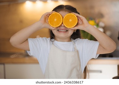 Close-up portrait of adorable little girl in apron stand in kitchen and smiling. Child holding halves of oranges at her eyes.  - Powered by Shutterstock