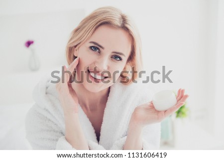 Closeup portrait of adult beautiful attractive blonde cute mature smiling woman wearing white bathrobe spreading face lotion cream gel on her face. Keeping and showing cream jar in other hand