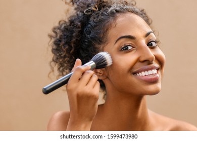 Closeup Portrait Of African Woman Applying Foundation With Makeup Brush. Black Beauty Girl Gets Blush On The Cheek Isolated On Light Brown Background. Portrait Of Young Woman Looking At Camera.