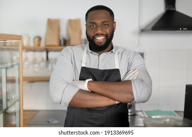 Close-up Portrait Of An African-American Waiter Standing With Arms Crossed In Cozy Loft Coffee Shop. A Confident Multiracial Small Business Owner Wearing Apron Looks At The Camera And Smiles