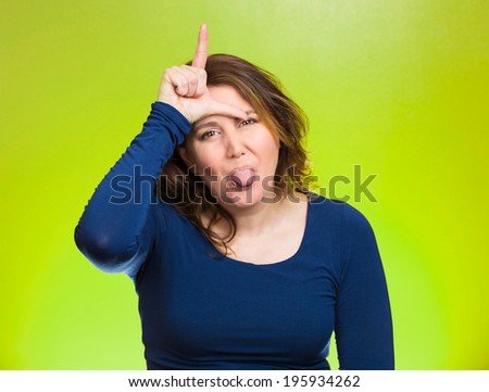 Closeup portrait angry, mad pissed woman, showing loser sign, sticking tongue out, hand on forehead, isolated green background. Negative emotion facial expression signs symbol feelings body language